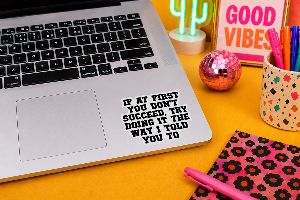 A silver laptop on a bright yellow desk with a black text decal that reads 'IF AT FIRST YOU DON'T SUCCEED, TRY DOING IT THE WAY I TOLD YOU TO'. Surrounding desk items include a pink notebook, a cup of pens, a neon cactus, and a 'GOOD VIBES' sign.