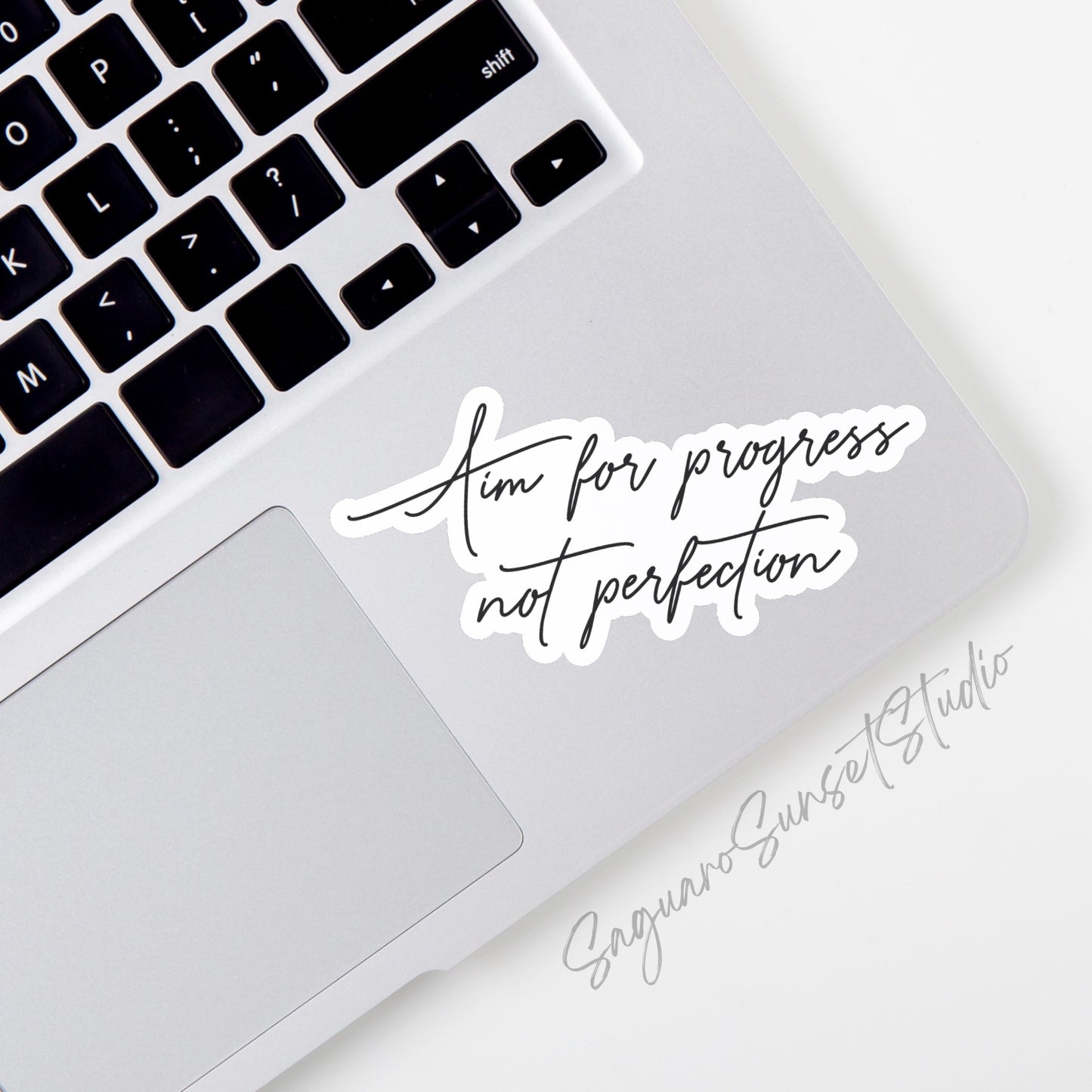 A motivational sticker on the corner of a laptop featuring the phrase 'Aim for Progress not Perfection' in a minimalist cursive design