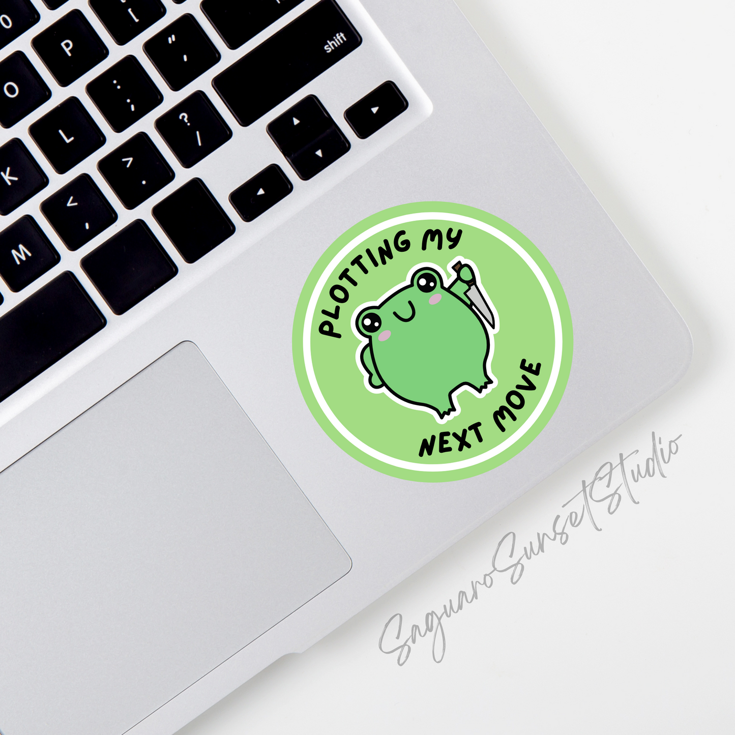 A green circle sticker featuring a mischievous frog holding a knife, with the text 'Plotting my next move' placed on the bottom corner of a laptop.