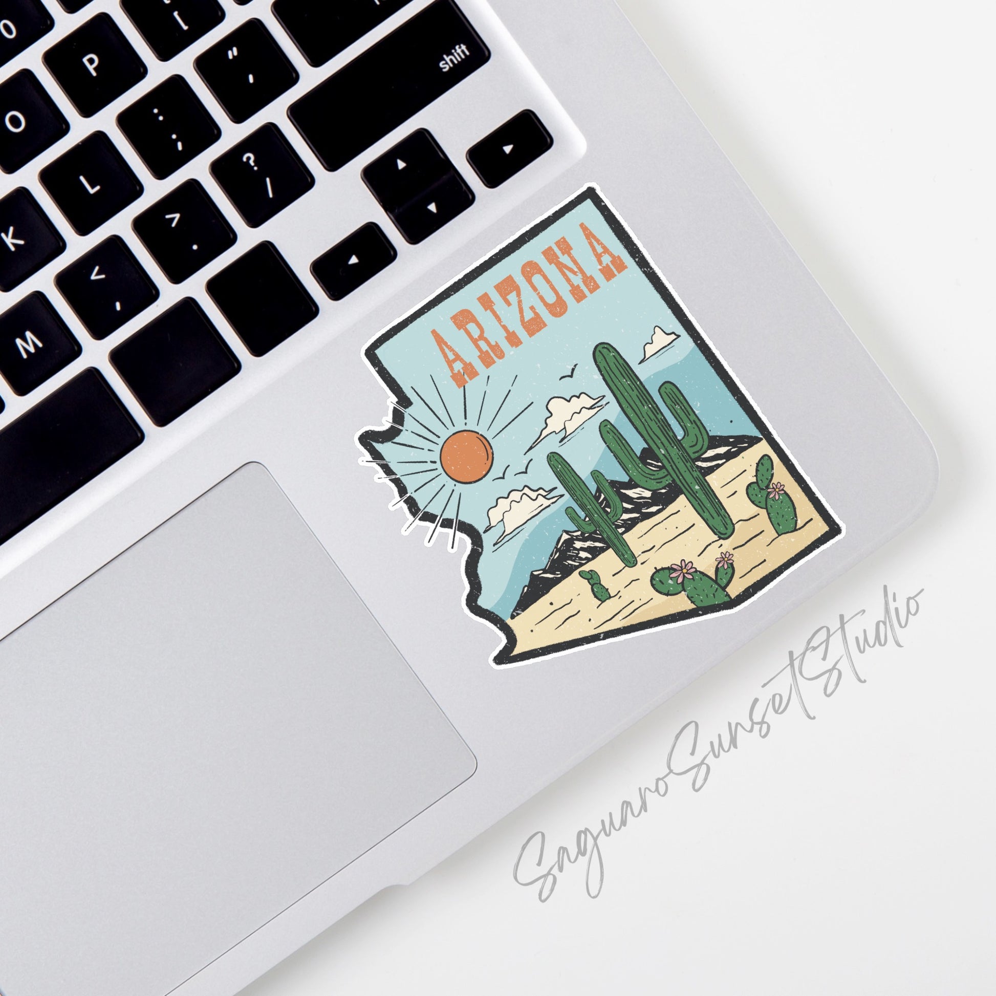 A sticker featuring the shape of Arizona filled with a desert landscape scene. The design includes tall cacti, blooming cacti, mountains, clouds, and a bright sun against a blue sky. The word "Arizona" is written in bold orange letters at the top. 