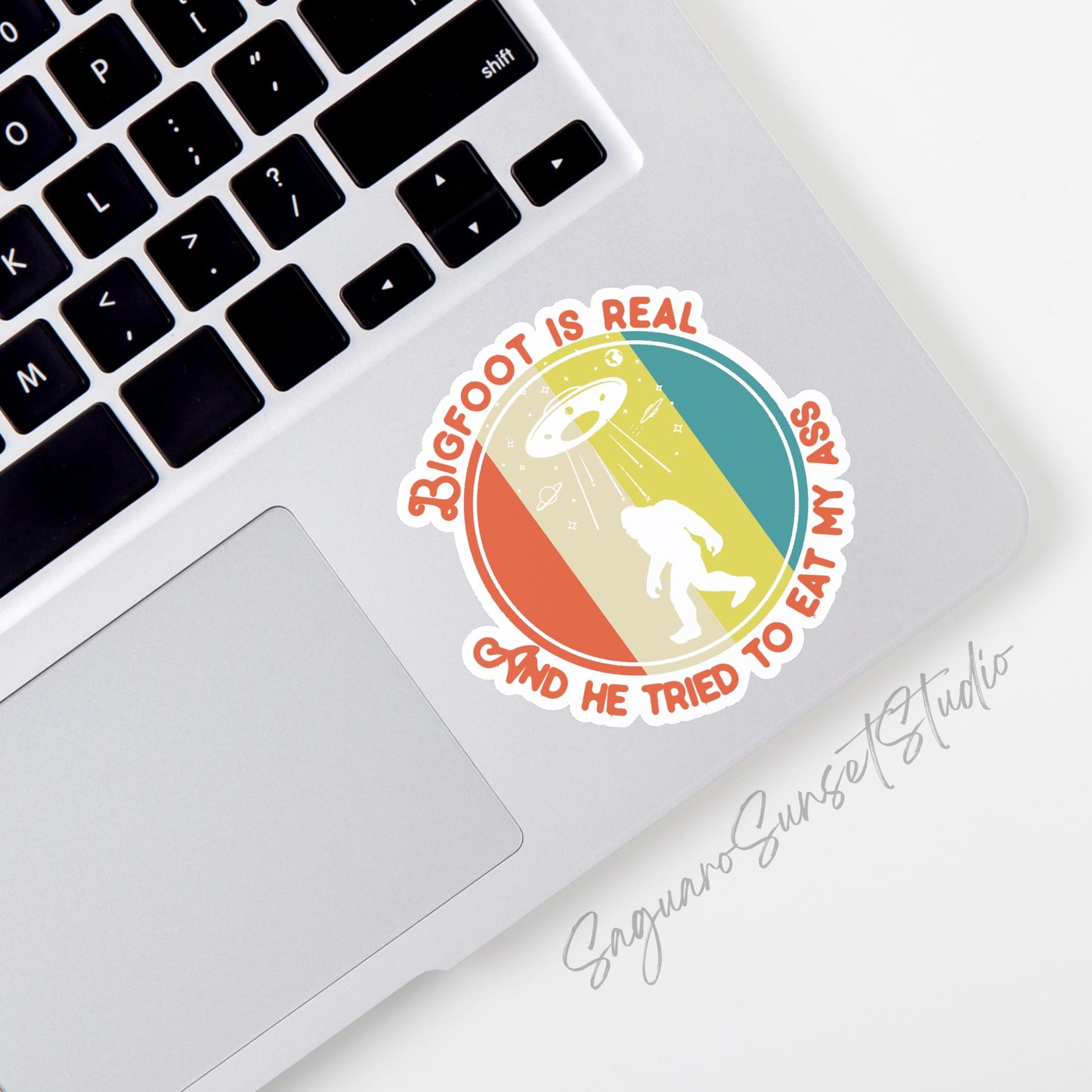 A sticker featuring the phrase "Bigfoot is Real and He Tried to Eat My Ass" written in retro-style orange text. The design includes a silhouette of Bigfoot with a UFO beaming down light in the background, set against a colorful striped backdrop. 