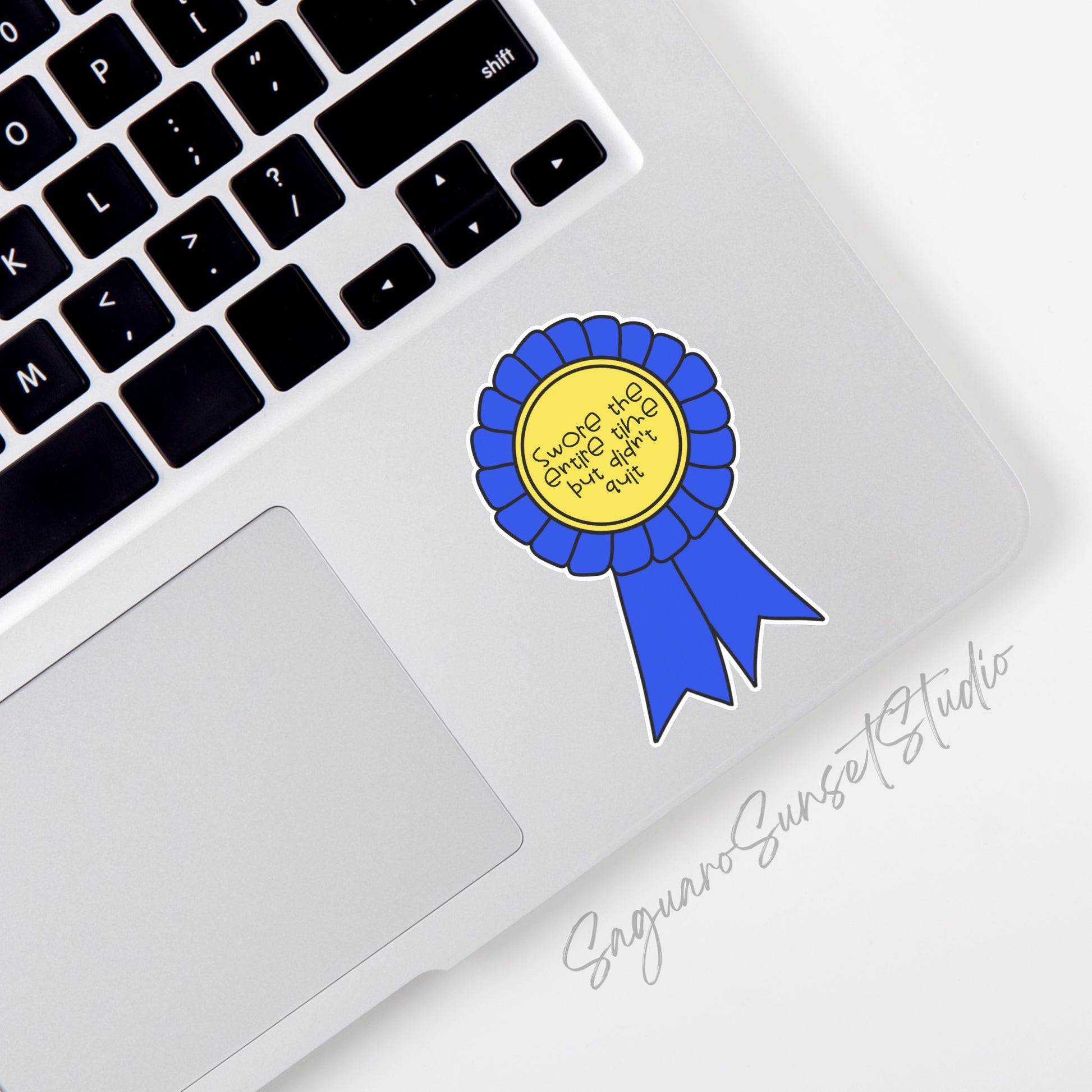 Sticker shaped like a blue ribbon with a yellow center, featuring the text "Swore the Entire Time But Didn't Quit," perfect for celebrating perseverance with a humorous twist.