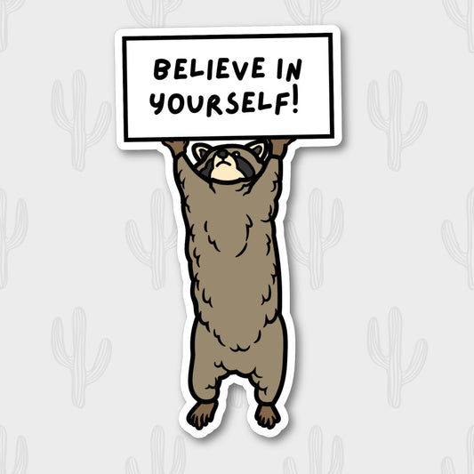 Sticker of a brown raccoon holding a white sign that says 'believe in yourself!'.