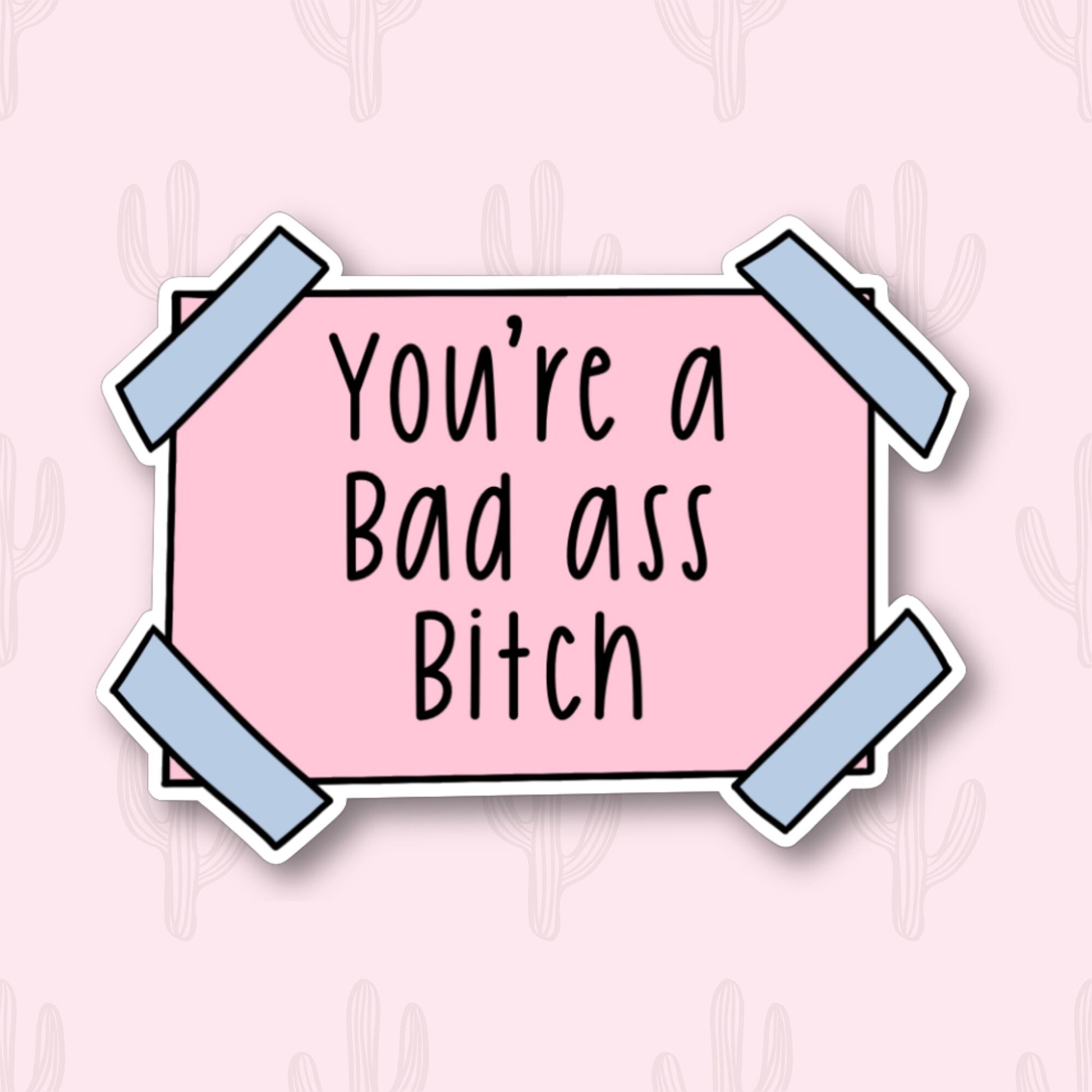 A sticker with the motivational phrase "You're a bad ass bitch" written in black text on a pink rectangular background. The sticker has blue tape corners.